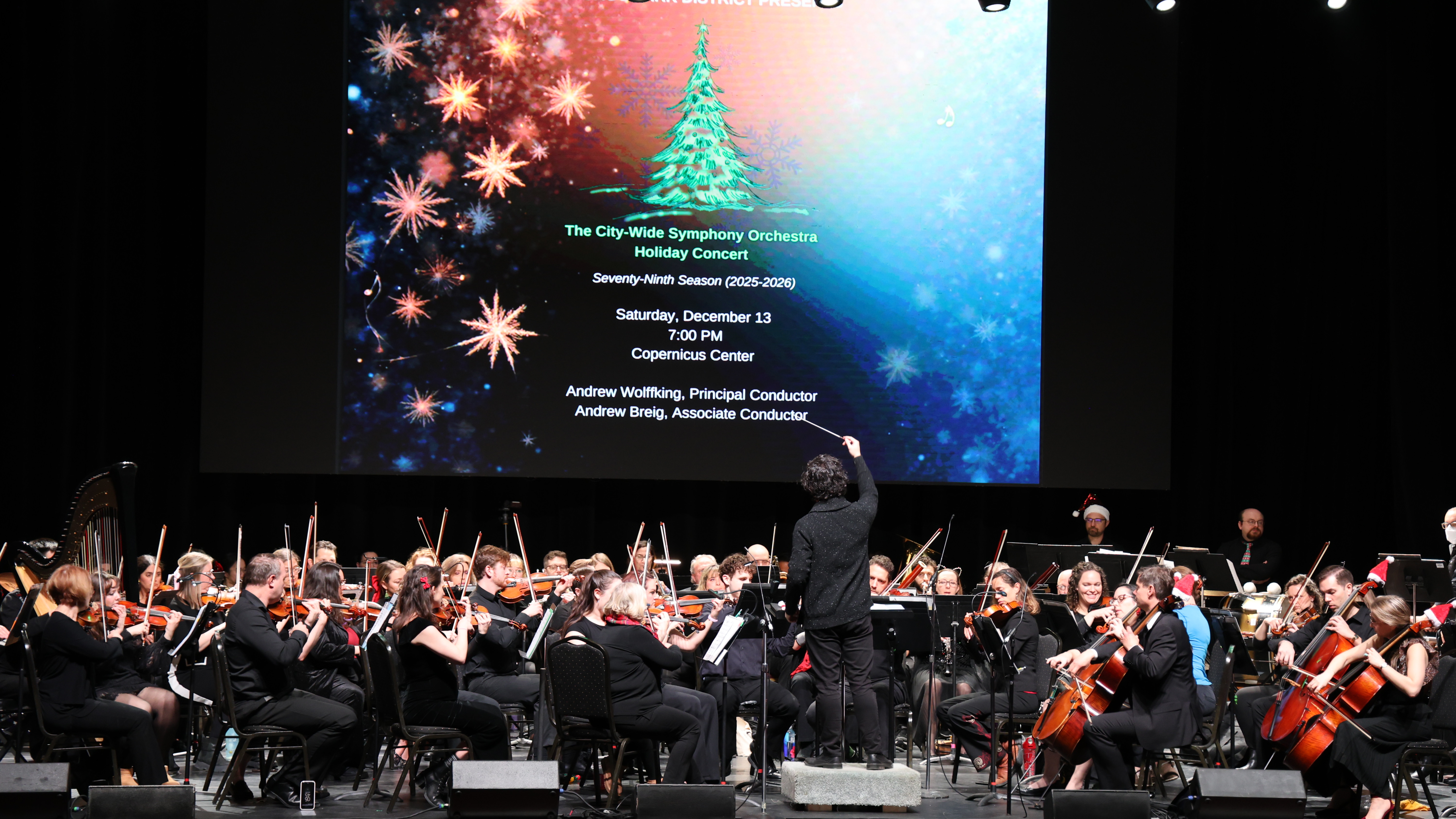 Orchestra plays with conductor in front of Christmas graphic.